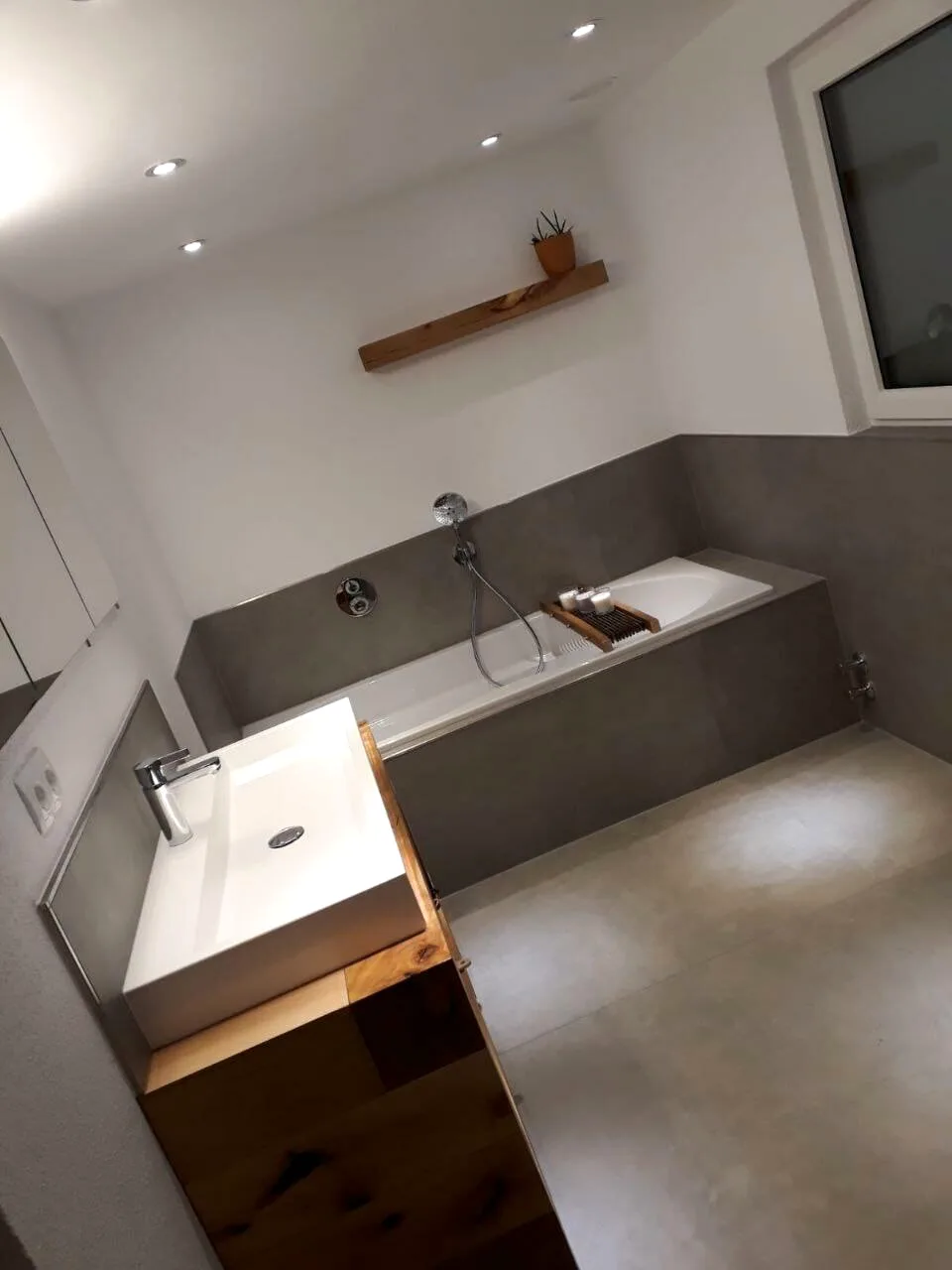 Badezimmer mit weißem Waschbecken auf Holzunterschrank, grauer Wanne und Wandregal