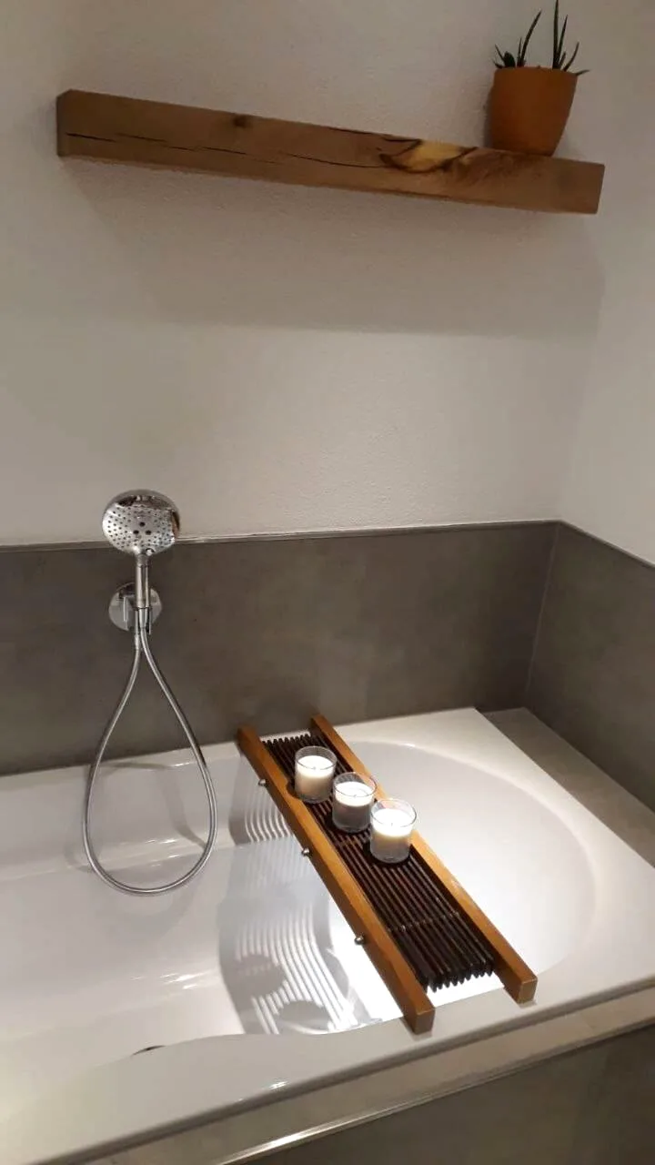 Badewanne mit Holzablage und Kerzen vor grauen Großformatfliesen, darüber ein Holzregal