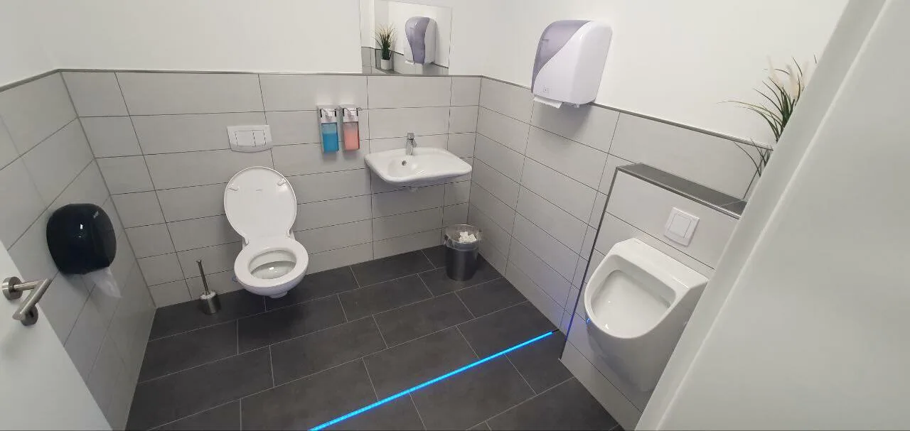 Sanitärraum für Gewerbe mit WC, Urinal, Waschbecken und grauen Fliesen